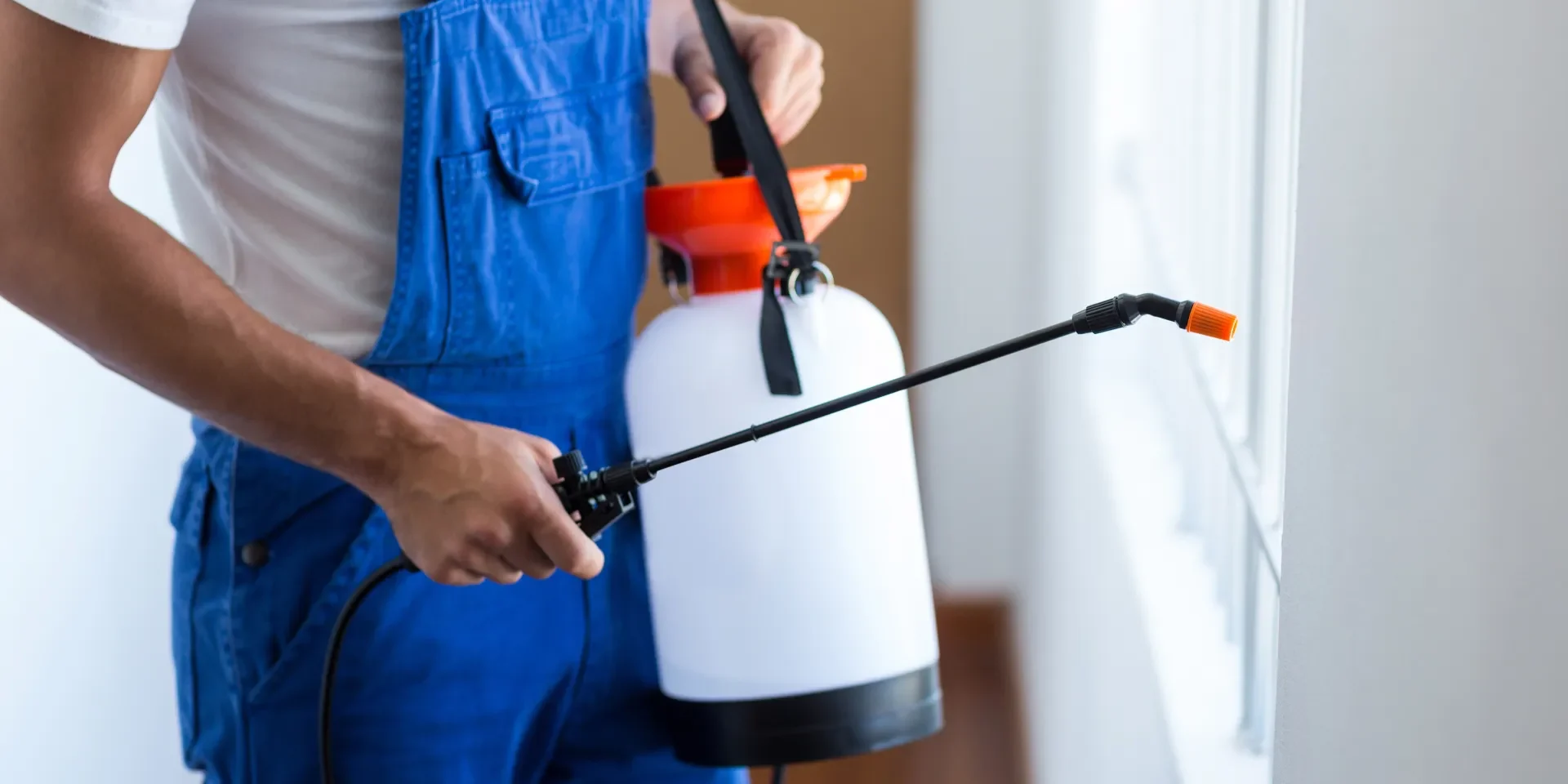 Pest control technician in blue overalls spraying pesticide along a window frame as part of general pest control services in Arizona